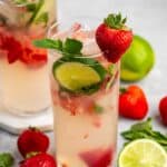 Two tall glasses of strawberry mojito sit on a gray surface. Each glass is garnished with sliced lime, mint leaves, and a whole strawberry. Fresh strawberries, lime slices, and mint leaves are scattered around the glasses.