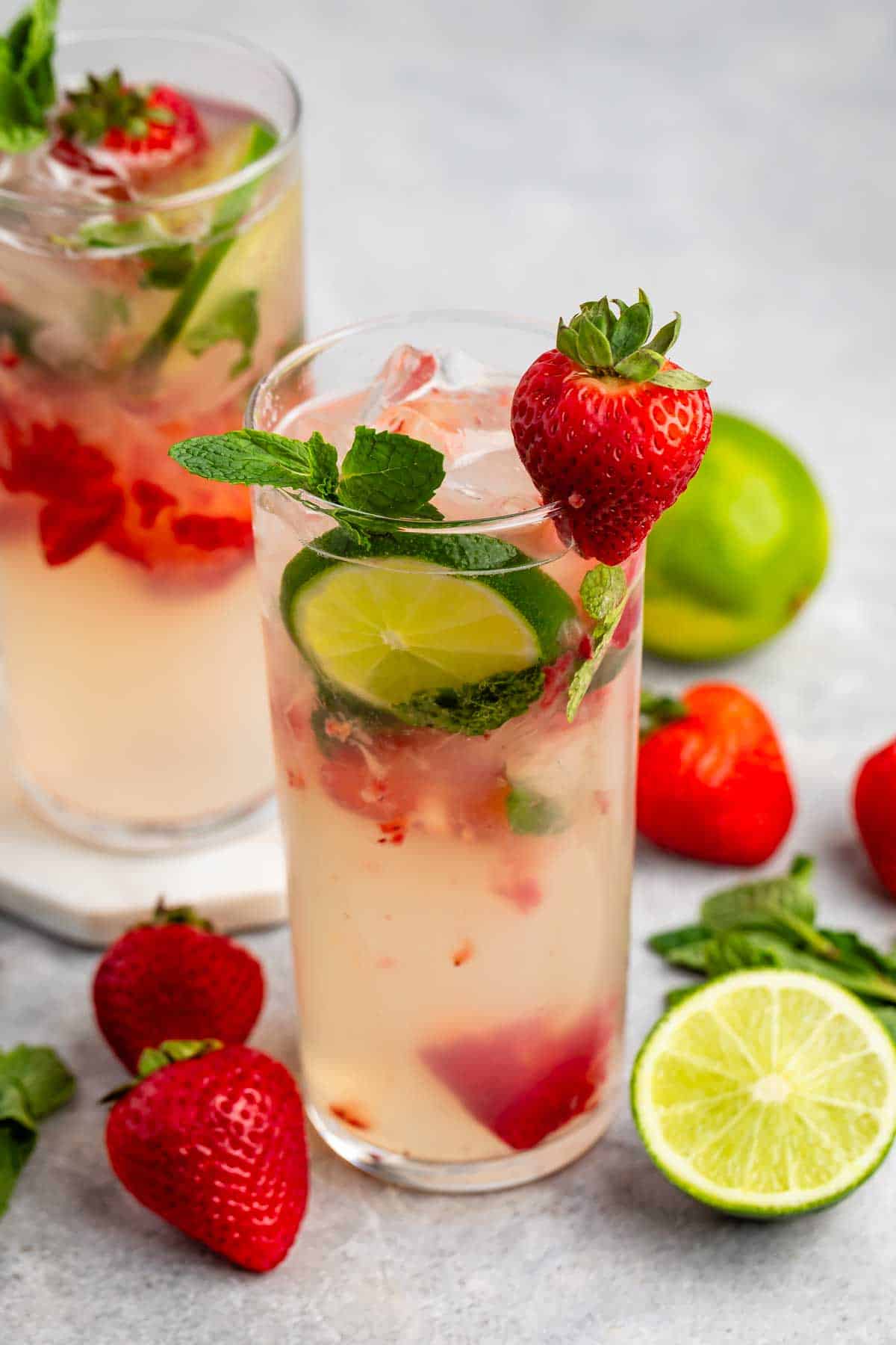 Two tall glasses of strawberry mojito sit on a gray surface. Each glass is garnished with sliced lime, mint leaves, and a whole strawberry. Fresh strawberries, lime slices, and mint leaves are scattered around the glasses.