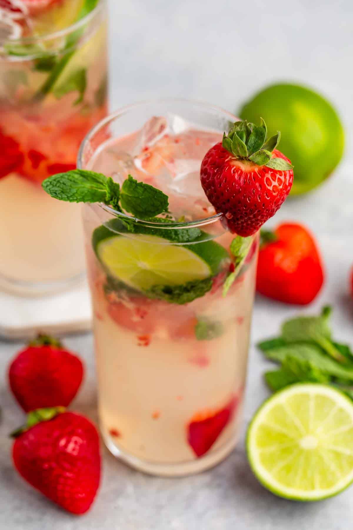 A refreshing drink in a clear glass filled with ice, lime slices, strawberries, and mint leaves. A whole strawberry garnishes the rim. Fresh strawberries and halved limes are scattered on the surface nearby.