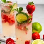 Two tall glasses of strawberry mojito garnished with strawberry, lime slices, and mint. Fresh strawberries, a lime half, and mint leaves are scattered around on a light surface.