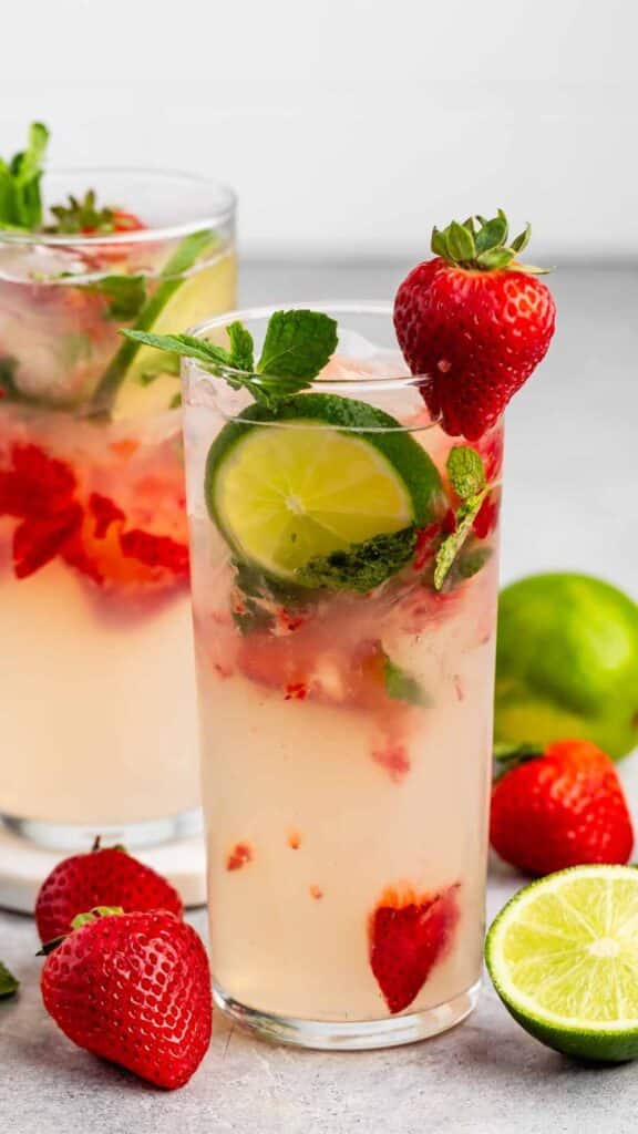 Two glasses of strawberry mojito garnished with lime slices, mint leaves, and strawberries. Fresh strawberries and a halved lime are scattered around the glasses on a gray surface.