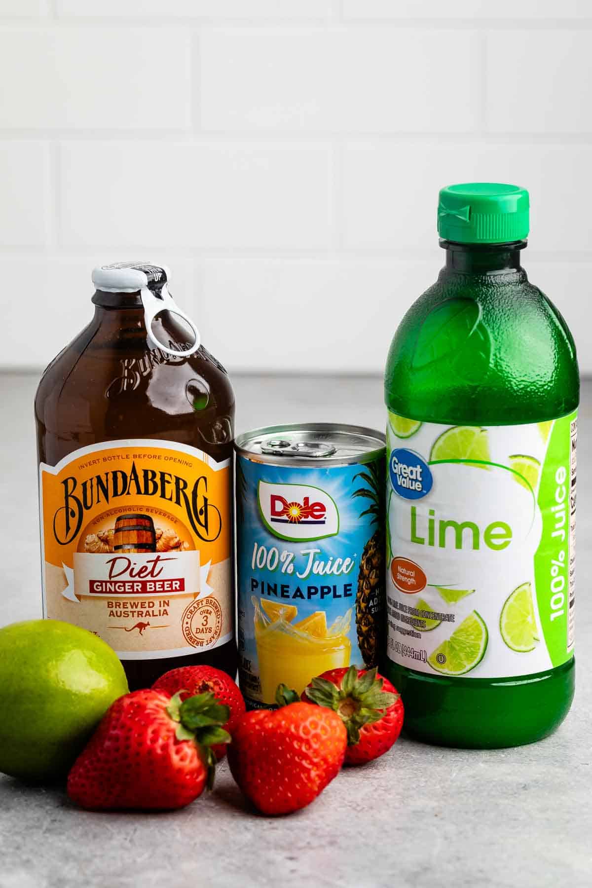 A selection of drink ingredients on a countertop: a bottle of Bundaberg Diet Ginger Beer, a can of Dole 100% Pineapple Juice, a bottle of Great Value Lime Juice, a fresh lime, and several strawberries.