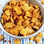 A bowl filled with a party mix of various cereal pieces, pretzels, and crunchy snacks, all seasoned, placed on a blue and white striped cloth. A few snack pieces are scattered around the bowl.