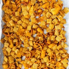 A baking sheet filled with a homemade snack mix. The mix includes various cereal squares and crunchy pretzel twists, all coated in a golden seasoning. The sheet is lined with white parchment paper.