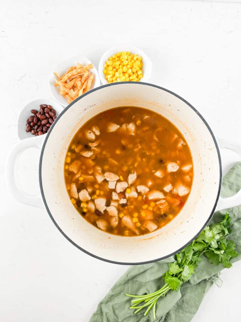 A pot of chicken tortilla soup with visible chunks of chicken, corn, and beans. Surrounding the pot are small bowls with tortilla strips, corn, and black beans. A bunch of cilantro and a green cloth are beside the pot on a white countertop.