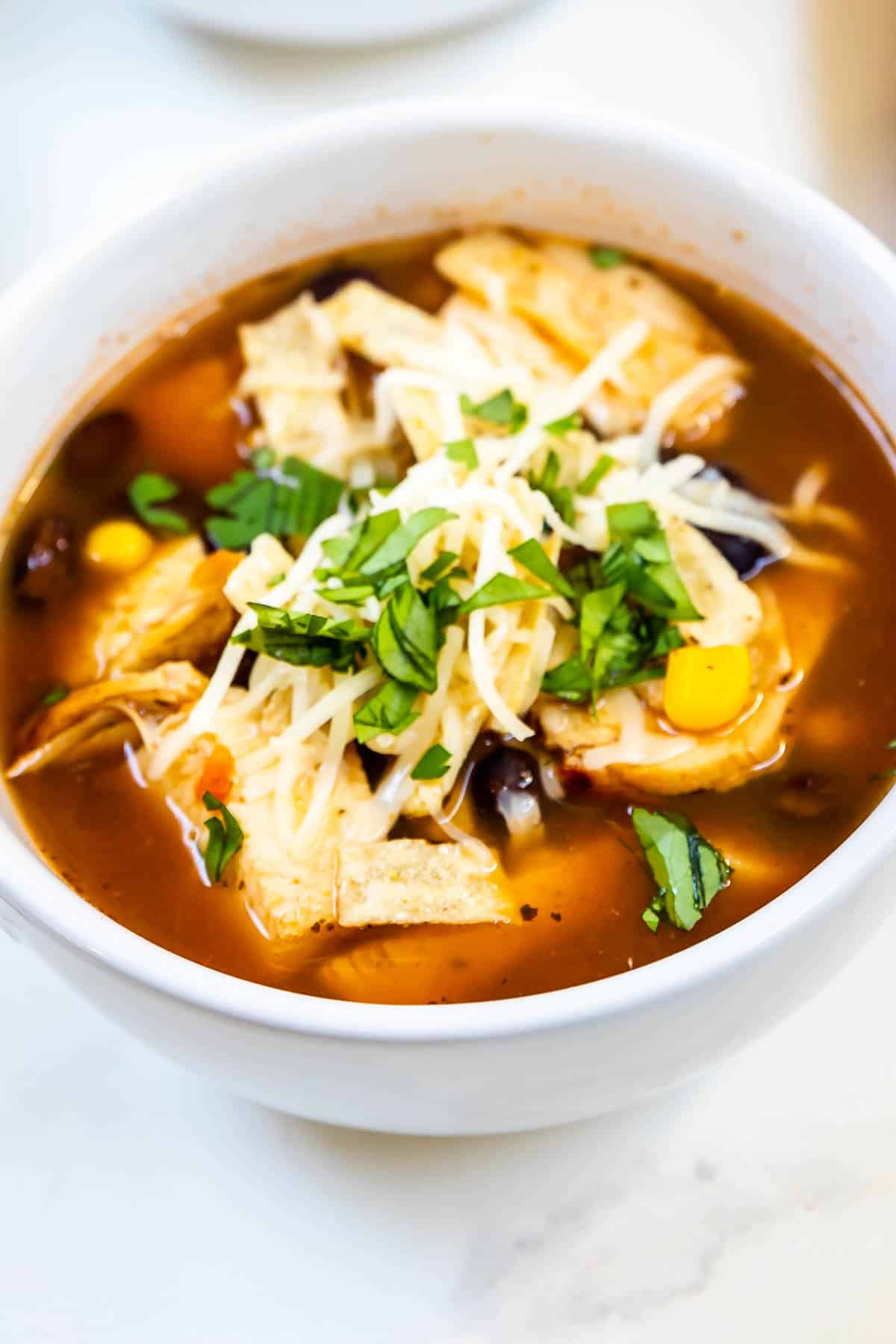 A bowl of tortilla soup topped with shredded cheese, tortilla strips, and chopped herbs. The soup contains black beans, corn, and pieces of chicken in a tomato-based broth. The bowl is on a white surface.