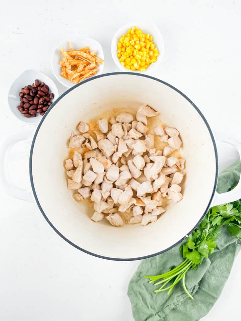 A pot filled with diced cooked chicken sits on a white surface. Surrounding the pot are small bowls with black beans, corn, and tortilla strips. A bunch of fresh cilantro is placed nearby on a green cloth.