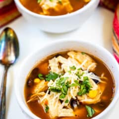 Two bowls of chicken tortilla soup topped with shredded cheese and fresh cilantro sit on a table. The soup includes visible pieces of chicken, corn, and garnished tortilla strips. A spoon and a red-striped cloth are nearby.