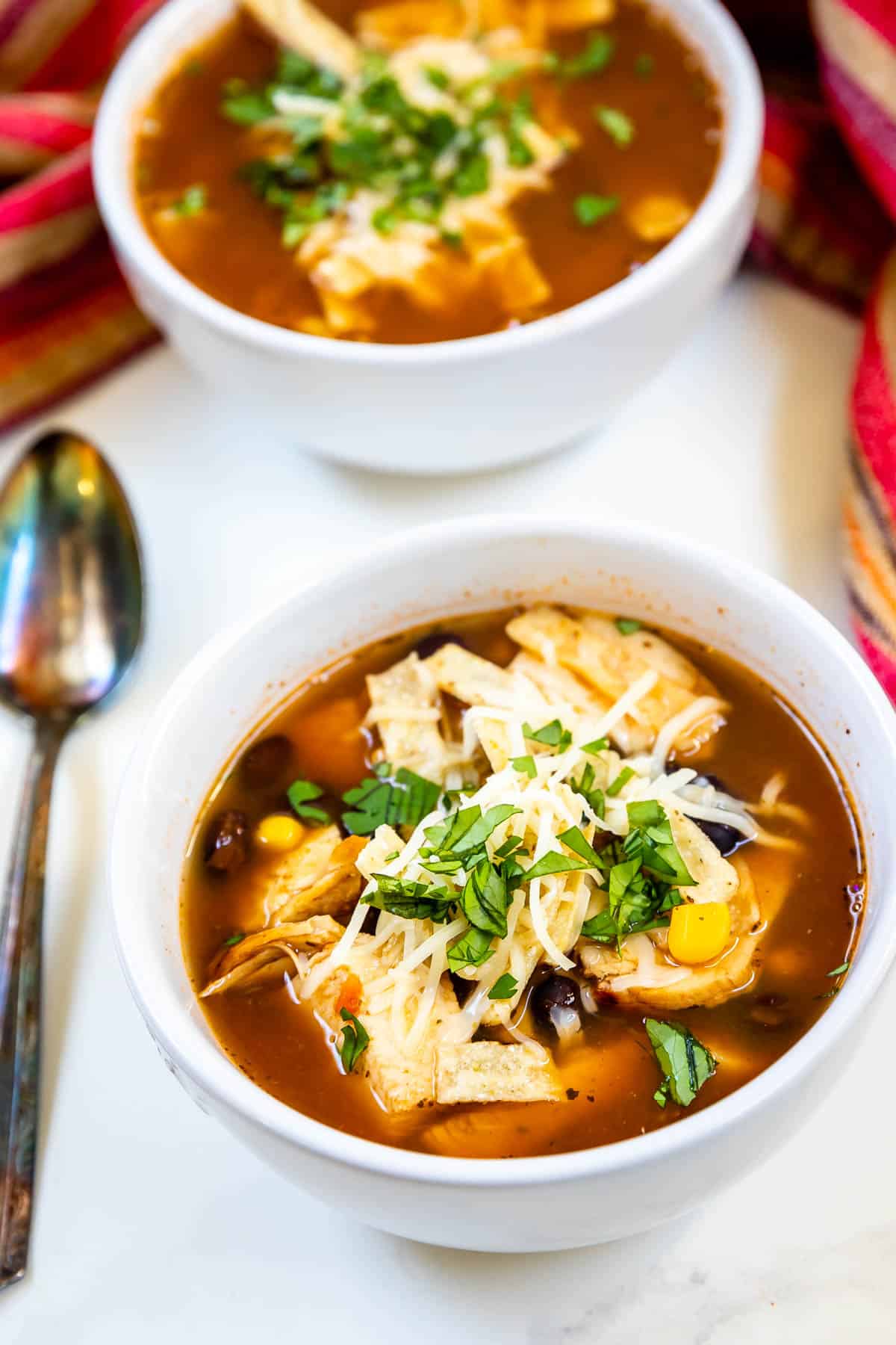 Two bowls of chicken tortilla soup topped with shredded cheese and fresh cilantro sit on a table. The soup includes visible pieces of chicken, corn, and garnished tortilla strips. A spoon and a red-striped cloth are nearby.