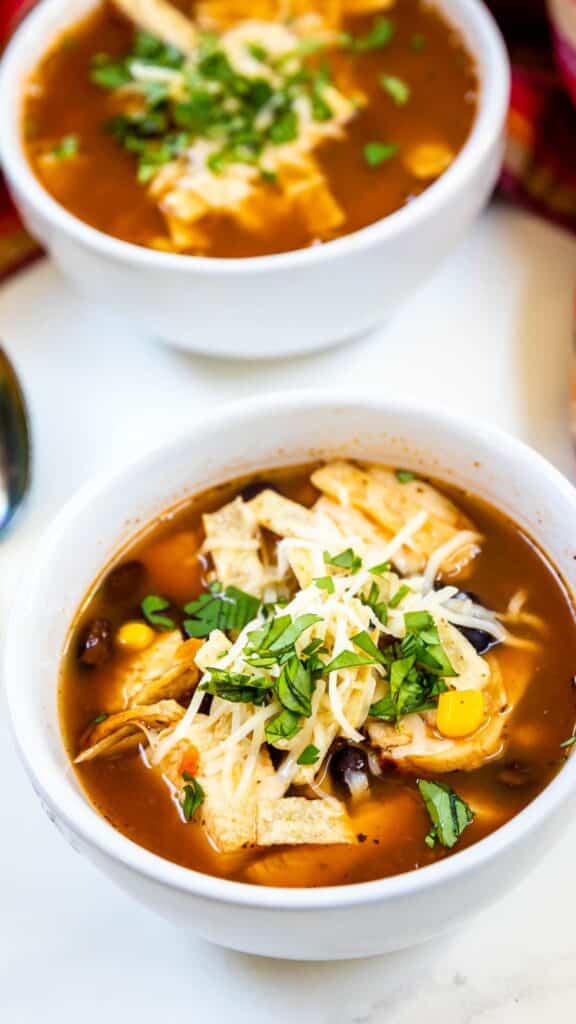 Two bowls of chicken tortilla soup topped with shredded cheese and fresh cilantro. The soup contains corn and black beans, and is served in white bowls on a light surface, with a hint of a spoon visible on the side.