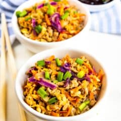 Two white bowls filled with a colorful mixture of ground meat, shredded carrots, purple cabbage, and sliced green onions. A small bowl of dark sauce and a pair of wooden chopsticks are placed nearby on a striped cloth.