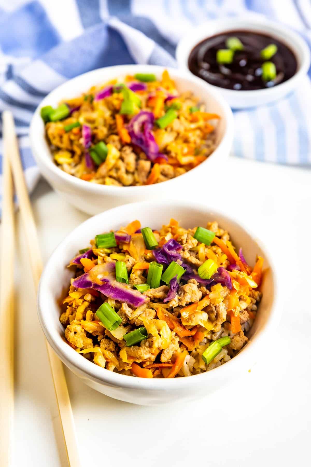 Two white bowls filled with a colorful mixture of ground meat, shredded carrots, purple cabbage, and sliced green onions. A small bowl of dark sauce and a pair of wooden chopsticks are placed nearby on a striped cloth.