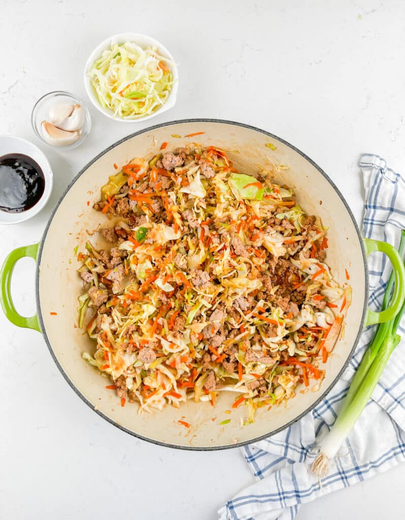 A large pot filled with a colorful mixture of chopped cabbage, carrots, and cooked meat. Nearby are small bowls of soy sauce, garlic cloves, and more cabbage. A green onion and a checkered cloth are placed on the white surface.