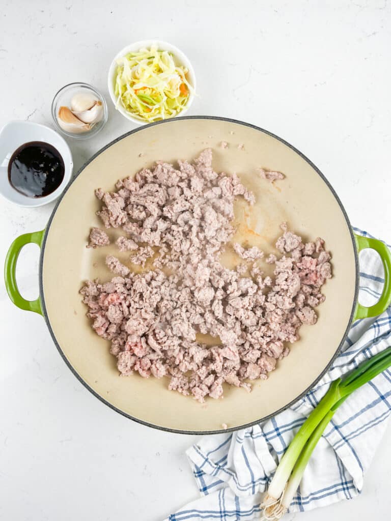 Overhead view of cooked ground meat in a large green-handled skillet. Nearby are small bowls with sauce, garlic, and shredded cabbage. A striped cloth and green onions are placed beside the skillet on a white countertop.