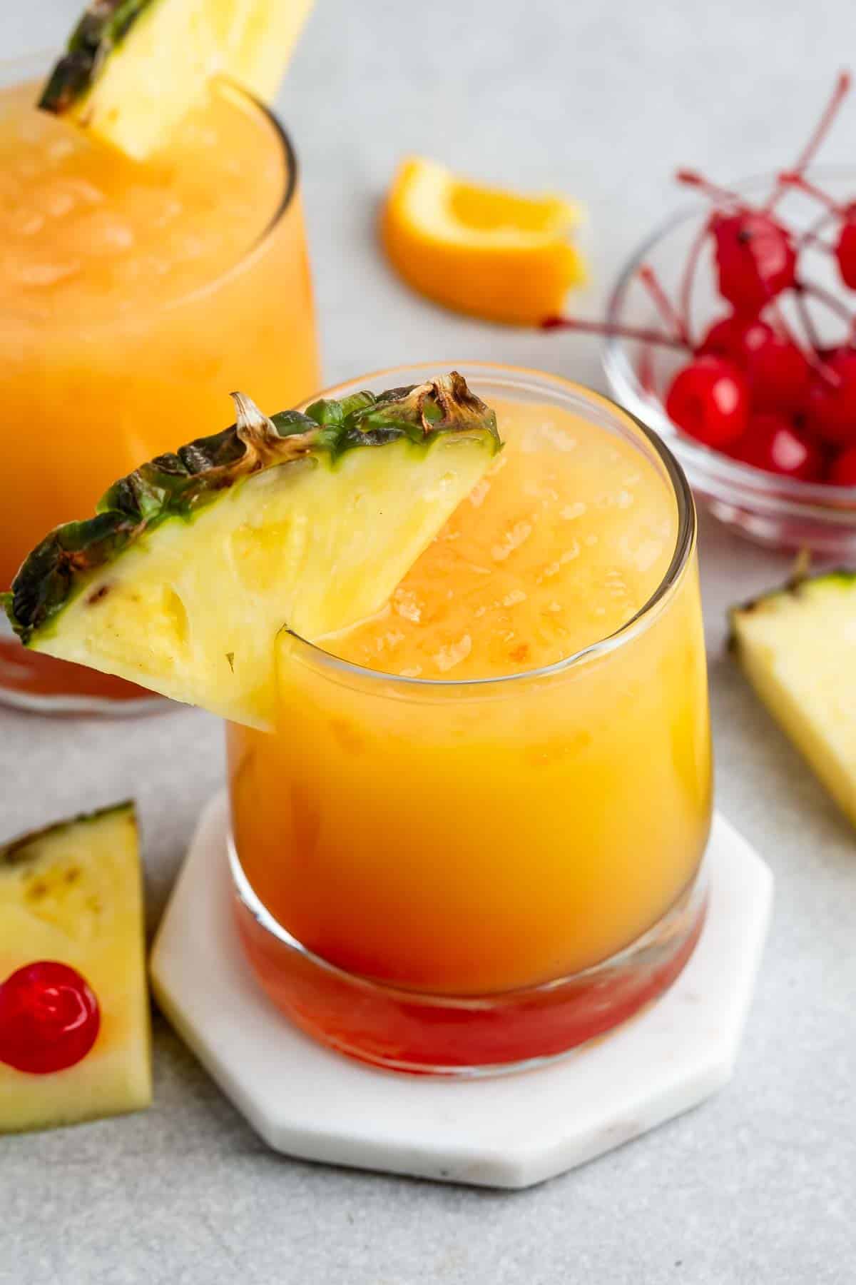 A vibrant tropical cocktail in a glass with a slice of pineapple garnish. The layered drink showcases hues of orange and red. Ice cubes float on top, with a background of cherries, pineapple slices, and orange wedges on a light surface.