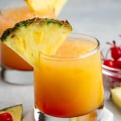 A glass of orange tropical cocktail garnished with a pineapple slice, with another similar drink in the background. A small dish with cherries and a pineapple slice are on the side. The surface is gray and the setting looks refreshing.