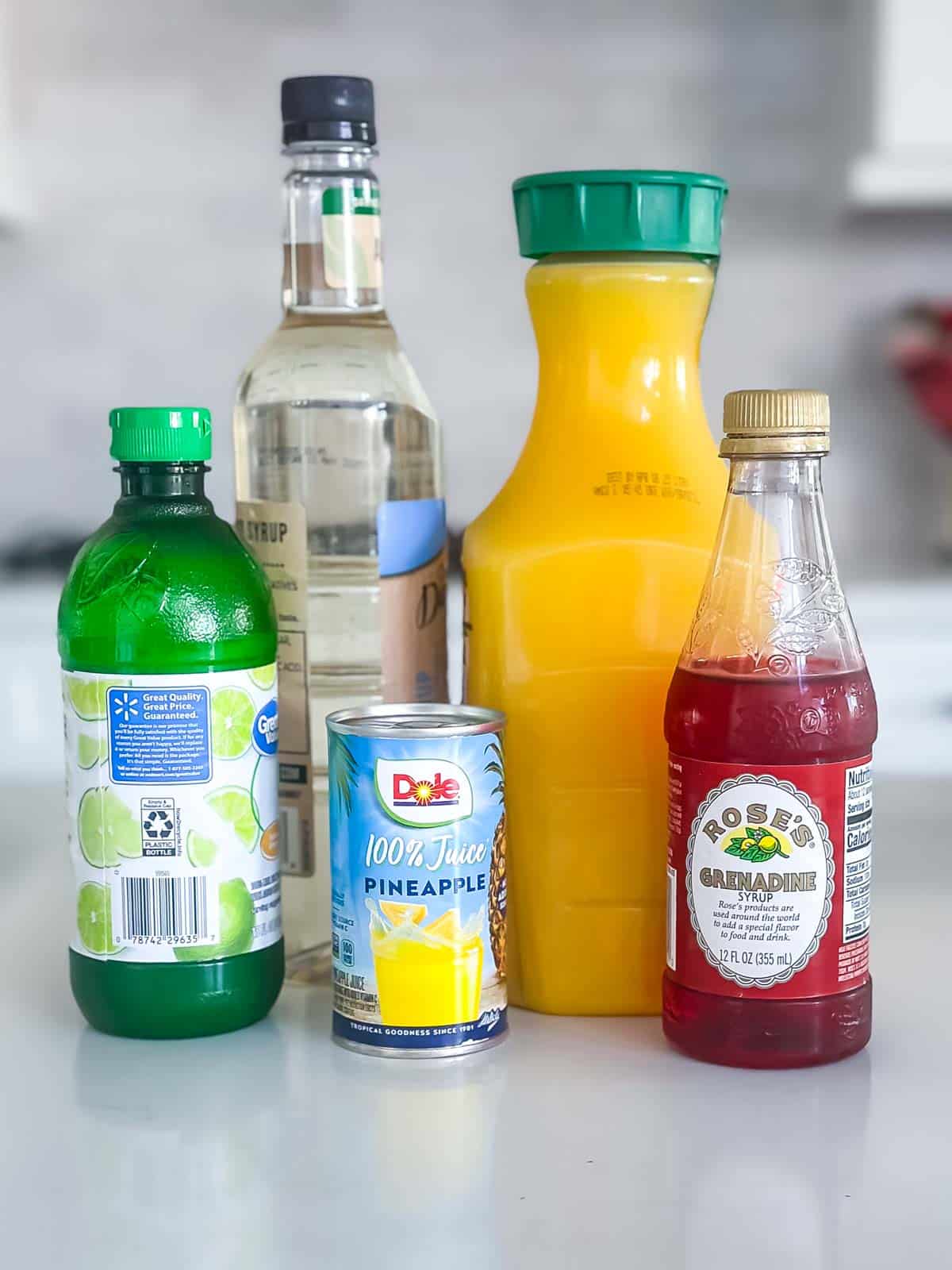 A variety of drink ingredients are displayed on a counter: a bottle of lime juice, a bottle of clear syrup, a bottle of white tequila, a large bottle of orange juice, a can of pineapple juice, and a bottle of grenadine syrup.