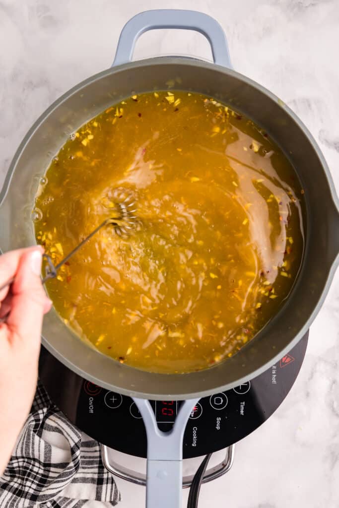 Hand stirring golden broth in a blue pot on a black induction cooktop. A whisk is used, and a checkered cloth is partially visible to the side.