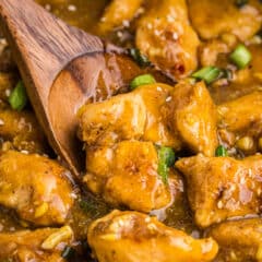 Close-up of a wooden spoon stirring chunks of orange chicken with a glossy sauce, garnished with sliced green onions.