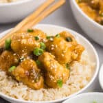 A dish of golden-brown battered chicken pieces garnished with sesame seeds and green onions sits atop a bed of white rice in a white bowl. Chopsticks rest on the bowls edge. Nearby are a bowl of plain rice and a small bowl of chopped green onions.