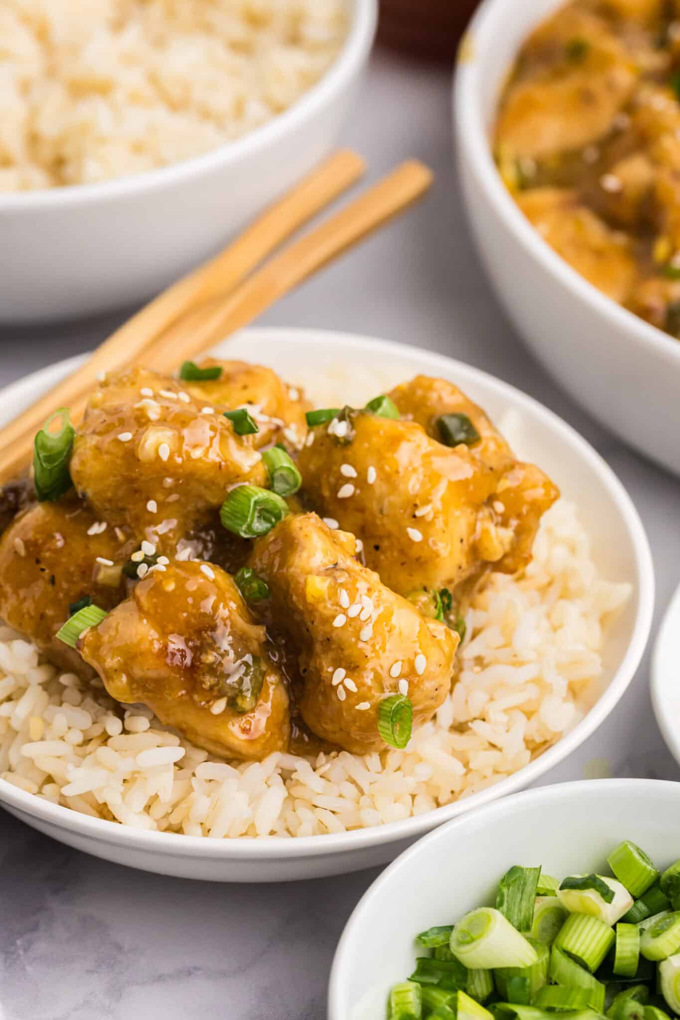 A dish of golden-brown battered chicken pieces garnished with sesame seeds and green onions sits atop a bed of white rice in a white bowl. Chopsticks rest on the bowls edge. Nearby are a bowl of plain rice and a small bowl of chopped green onions.