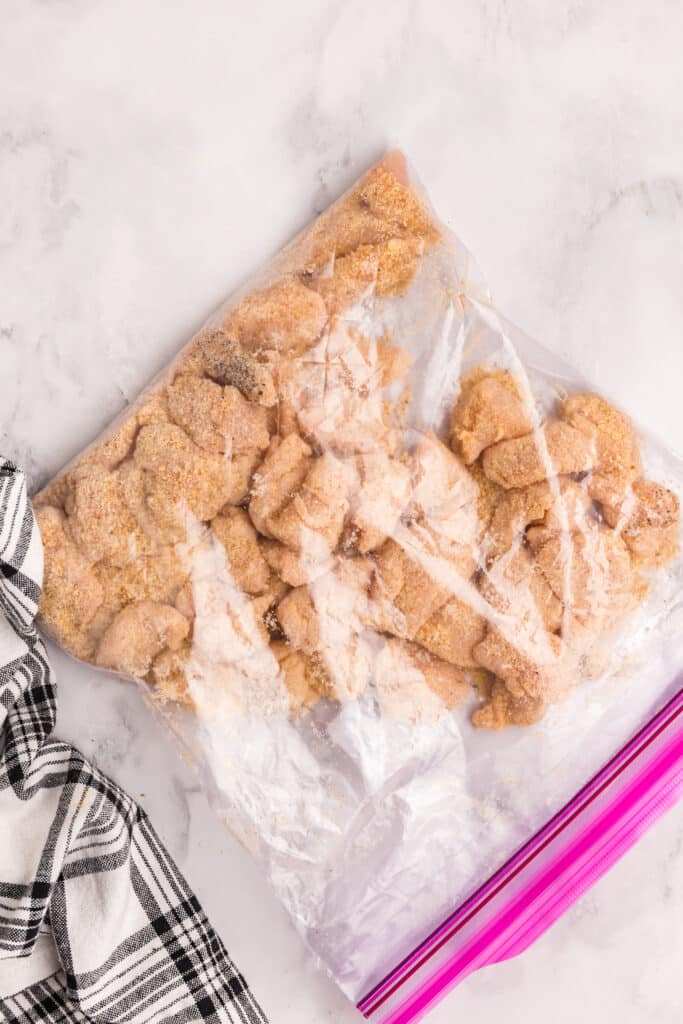 A clear plastic ziplock bag filled with breaded chicken pieces resting on a white marble surface. A black and white plaid towel is partially visible on the left side.