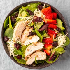 A bowl of fresh salad with sliced grilled chicken, strawberries, dried cranberries, shredded cheese, and sunflower seeds, served on a bed of mixed greens. The bowl sits on a textured gray surface.