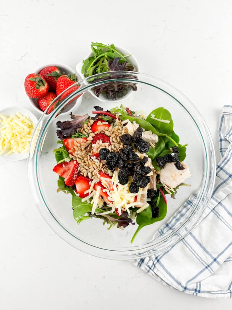 A glass bowl filled with a salad of mixed greens, sliced strawberries, chicken pieces, shredded cheese, sunflower seeds, and dried cranberries. Beside it are fresh strawberries, shredded cheese, and a small bowl of greens on a white surface.