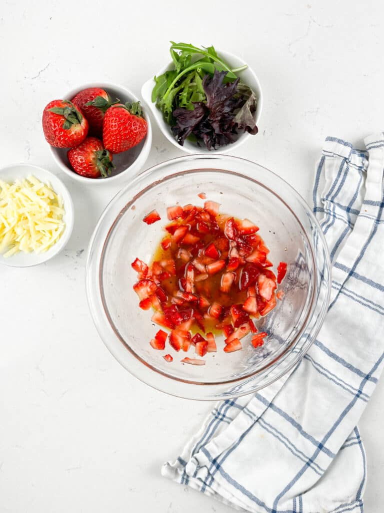 A clear bowl with diced tomatoes in liquid sits on a white surface. Surrounding it are small bowls with strawberries, shredded cheese, and mixed greens. A white and blue striped cloth is nearby.