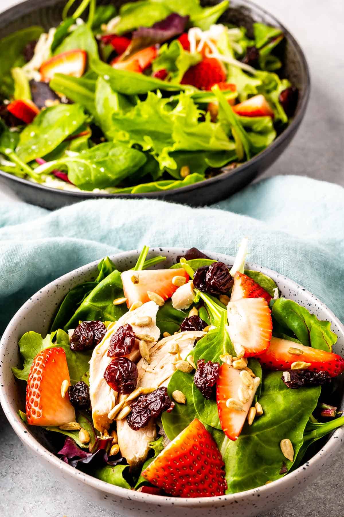 Two bowls of fresh salad featuring mixed greens, sliced strawberries, dried cranberries, sunflower seeds, and pieces of chicken, placed on a light blue cloth. The foreground bowl is in focus, with the background bowl slightly blurred.