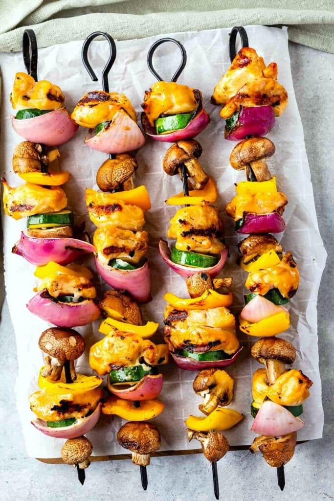 Skewers of grilled chicken, bell peppers, zucchini, red onions, and mushrooms, arranged on parchment paper. The vegetables and chicken are charred, showcasing a colorful and appetizing presentation.