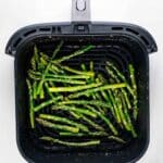Air fryer basket filled with cooked asparagus spears, lightly seasoned, on a white background. The asparagus is evenly distributed across the grill surface.