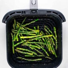 Air fryer basket filled with cooked asparagus spears, lightly seasoned, on a white background. The asparagus is evenly distributed across the grill surface.
