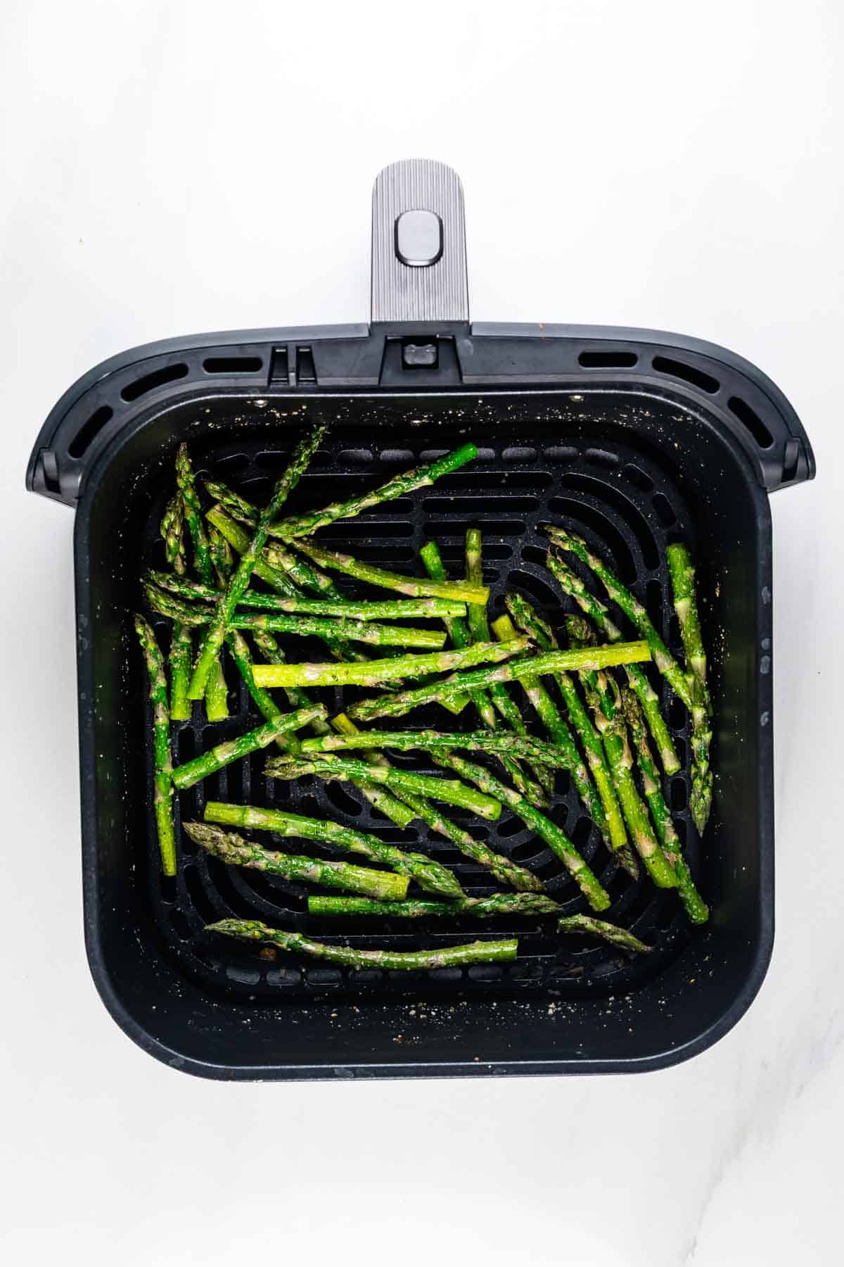 Air fryer basket filled with cooked asparagus spears, lightly seasoned, on a white background. The asparagus is evenly distributed across the grill surface.