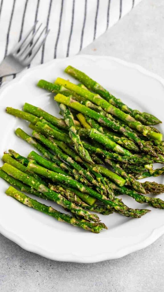 A white plate filled with roasted asparagus spears, seasoned with herbs and spices. A striped napkin and fork are partially visible in the background on a light surface.