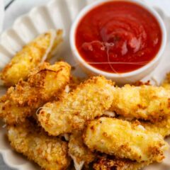 A plate of crispy, breaded mozzarella sticks is arranged neatly on a scalloped dish. A small bowl of marinara sauce accompanies the sticks, providing a classic dipping option. The setup is on a light surface.