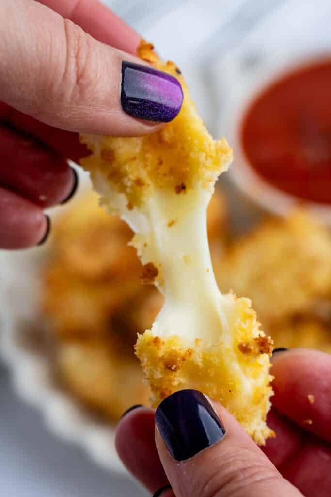 Close-up of hands pulling apart a breaded mozzarella stick, with melted cheese stretching between the halves. The person has dark purple nail polish. A bowl of marinara sauce is blurred in the background.