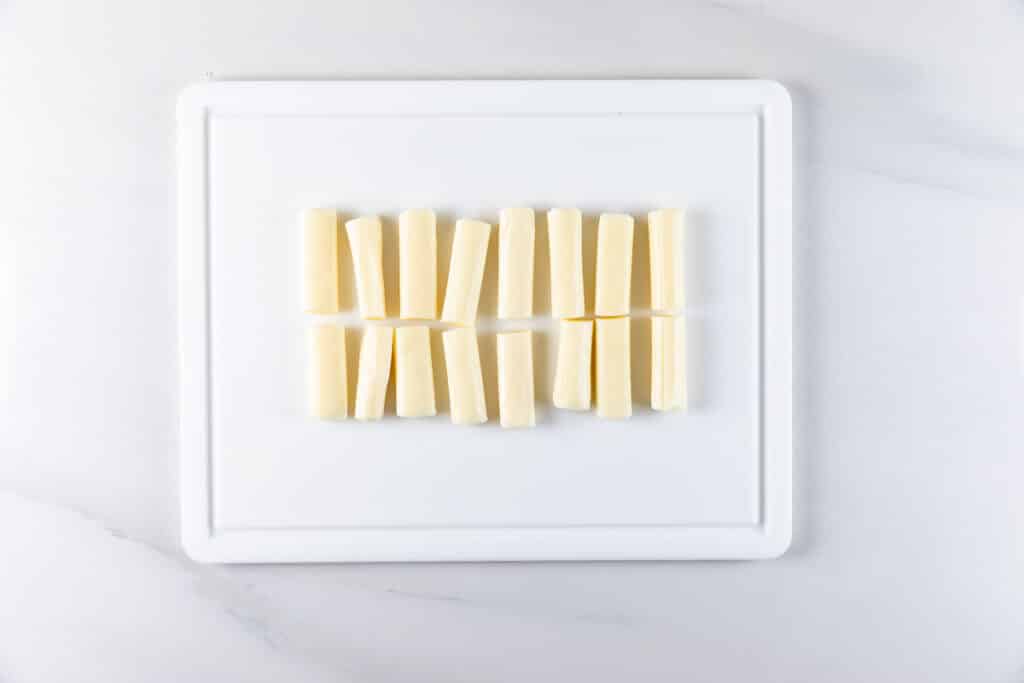 A white cutting board with evenly spaced sticks of string cheese, each cut in half, laid out in two parallel rows against a white background.