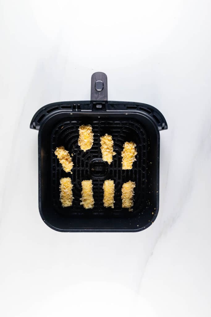 Seven breaded fish sticks evenly spaced in a black air fryer basket, viewed from above, on a white background.
