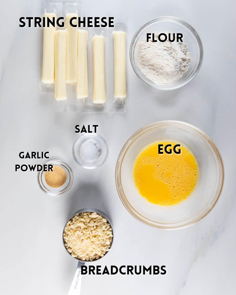 Ingredients for making mozzarella sticks on a marble surface: string cheese, flour, an egg in a bowl, breadcrumbs, garlic powder, and salt. Each item is in a separate bowl or container.