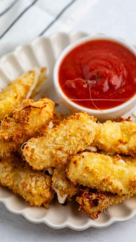 A plate filled with crispy mozzarella sticks, coated in golden breadcrumbs, is accompanied by a small bowl of marinara sauce. The plate is placed on a light-colored surface with a striped cloth in the background.