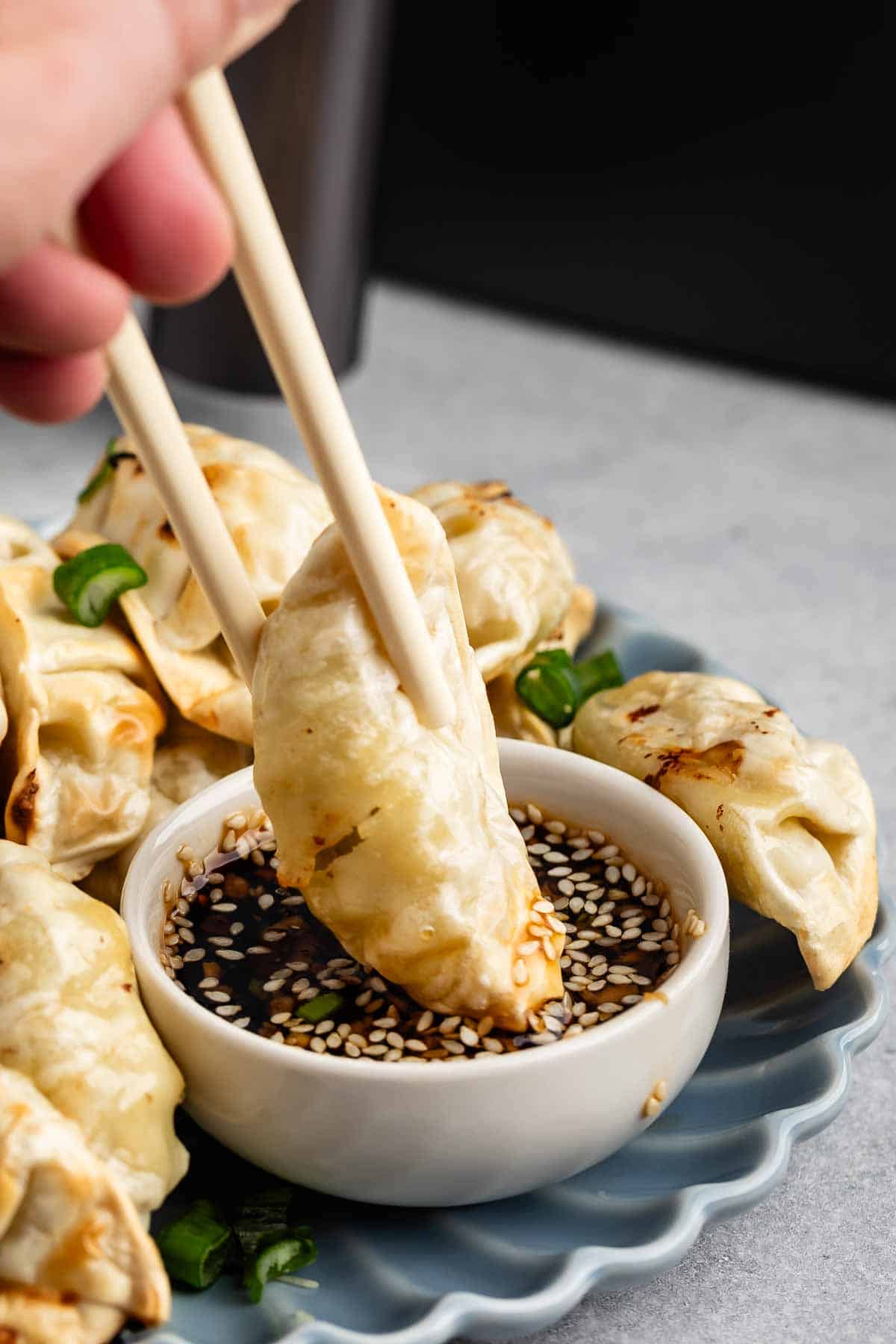 A hand uses chopsticks to hold a dumpling above a small bowl of soy sauce topped with sesame seeds and sliced green onions. More dumplings are arranged on a blue plate around the bowl.