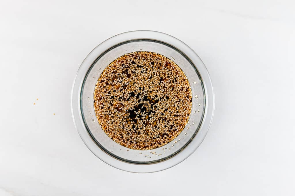 A clear bowl filled with soy sauce, sesame seeds, and chopped garlic on a white surface.