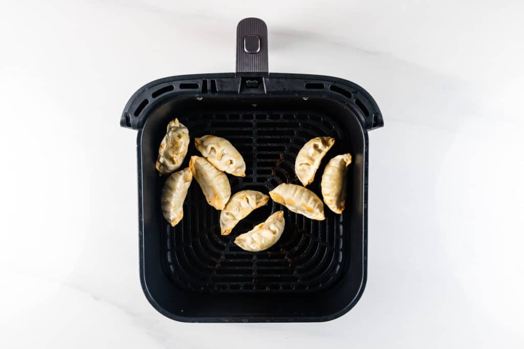 Top view of several golden-brown dumplings arranged inside an air fryer basket on a white surface. The dumplings are evenly spaced, showcasing their crispy, cooked appearance.
