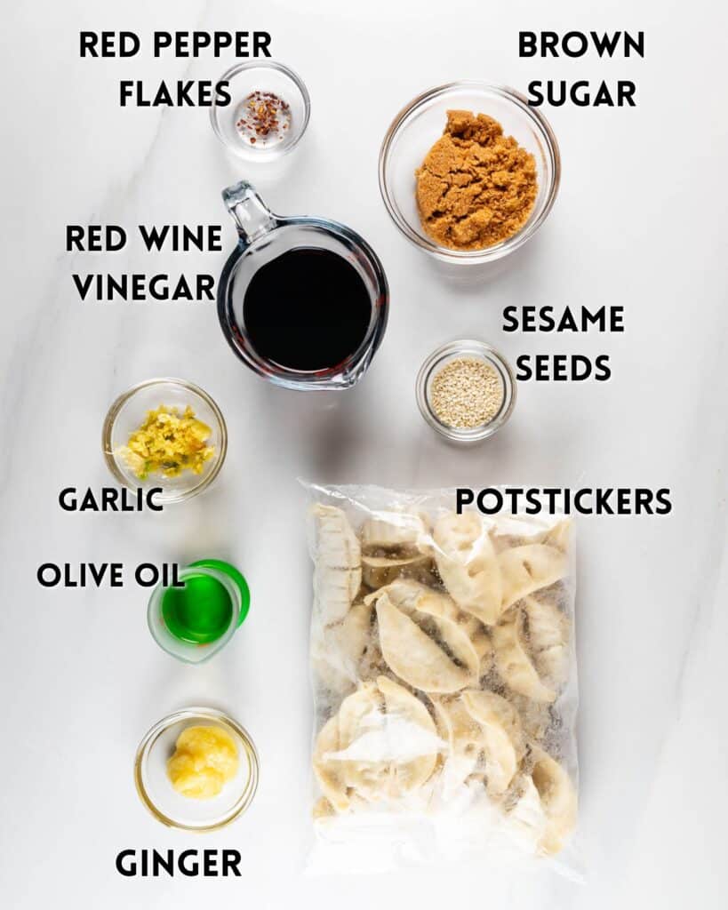 Ingredients for a potsticker dish arranged on a white surface: a bag of potstickers, small bowls of brown sugar, sesame seeds, minced garlic, and ginger, a cup of red wine vinegar, red pepper flakes, and a small bowl of olive oil.