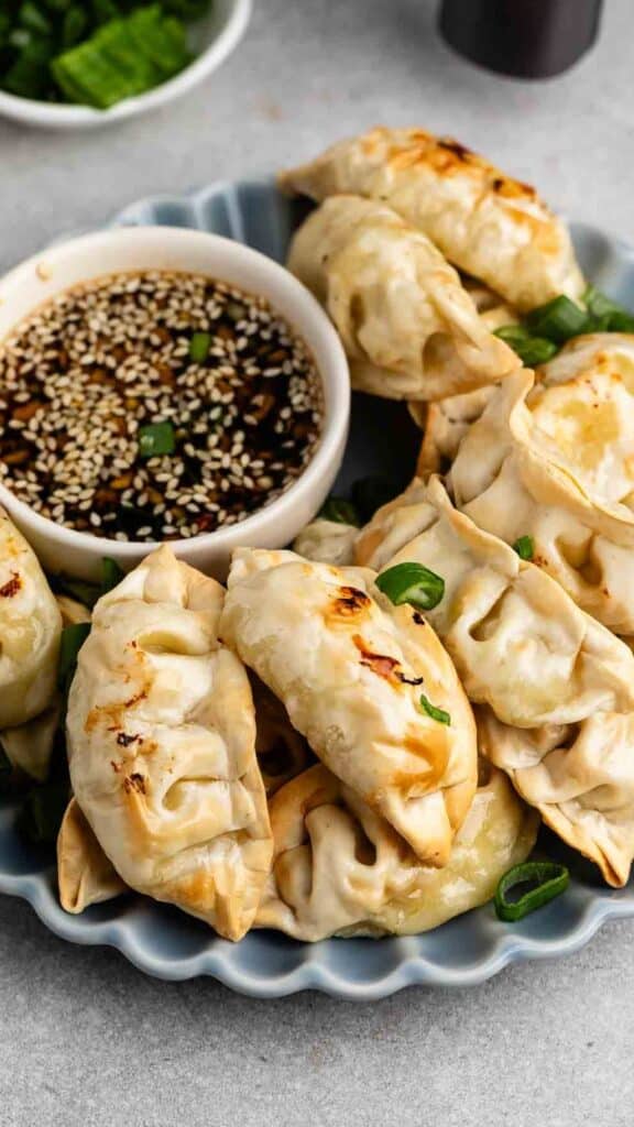 A plate filled with golden-brown dumplings garnished with green onions, accompanied by a small bowl of soy sauce topped with sesame seeds, sitting on a gray surface.