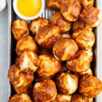 A tray filled with golden-brown pretzel bites, accompanied by a small dish of yellow mustard for dipping. The tray rests on a light-colored surface, with a partial view of a striped cloth in the corner.