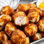 A tray filled with golden-brown pretzel bites, with a soft, fluffy interior visible in one. A small bowl of yellow mustard is visible in the background next to a striped cloth.