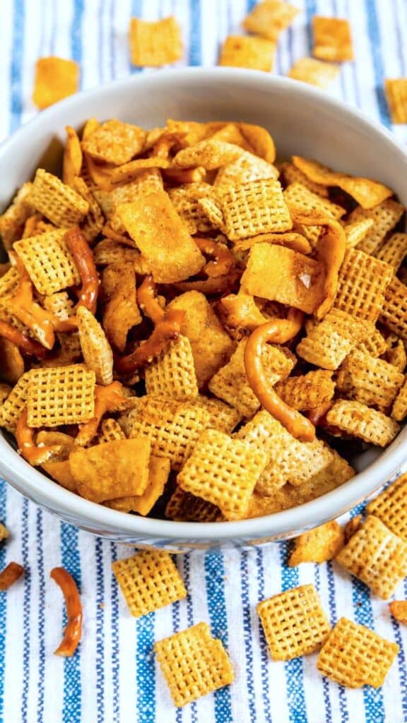 A bowl filled with a snack mix of Chex cereal, pretzels, and other crunchy pieces. The nuts and cereal are lightly seasoned. The bowl is placed on a striped blue and white cloth, with some snacks scattered around.