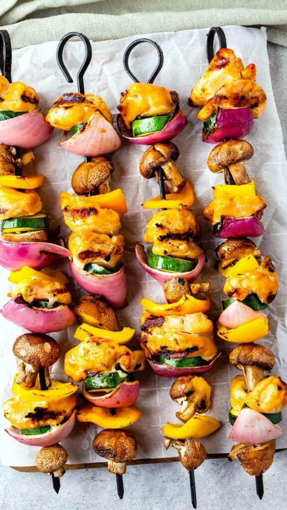 Skewers of grilled chicken, bell peppers, zucchini, red onions, and mushrooms, arranged on parchment paper. The vegetables and chicken are charred, showcasing a colorful and appetizing presentation.